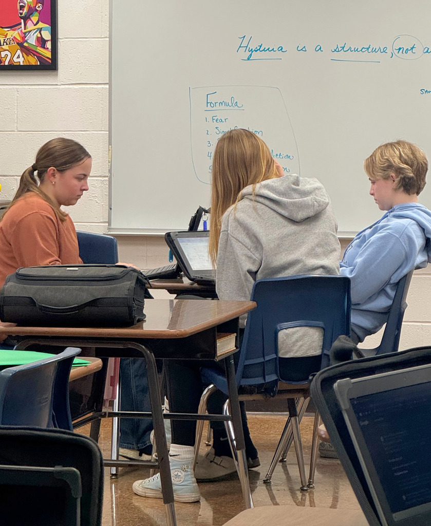 A small group of students are sitting around the desks. They are researching on their Chromebooks. and discussing. 