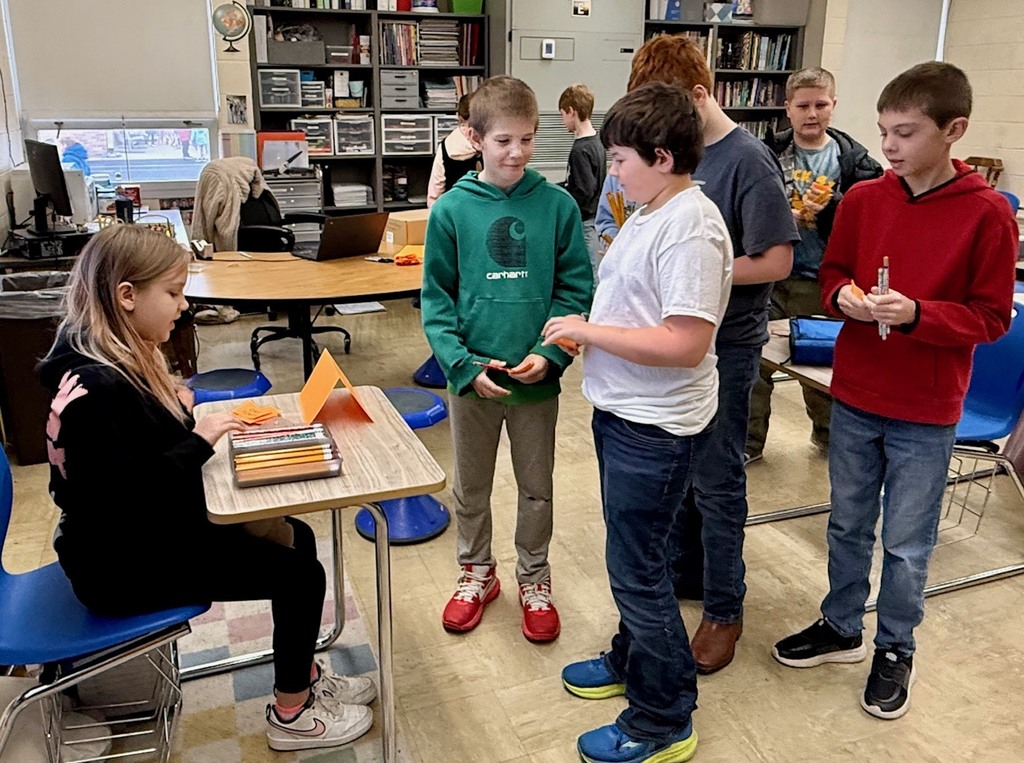 A student is at another student's desk buying a pencil. Other students are looking on.