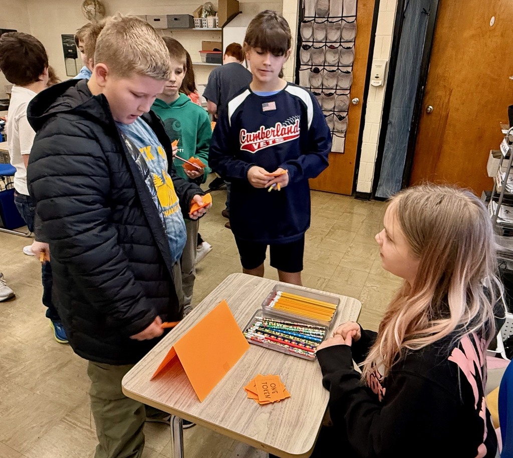A student is at another student's desk buying a pencil. Other students are looking on.