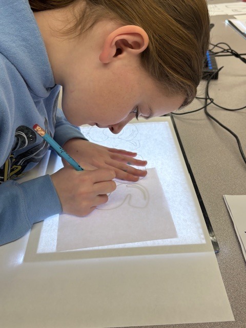Students are tracing bubble letters using a light board. Their paper and the letter they are tracing on top of a board that is electronically lit up.