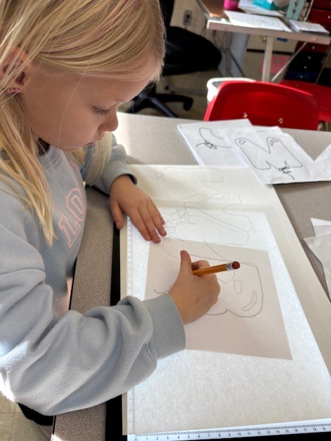Students are tracing bubble letters using a light board. Their paper and the letter they are tracing on top of a board that is electronically lit up.