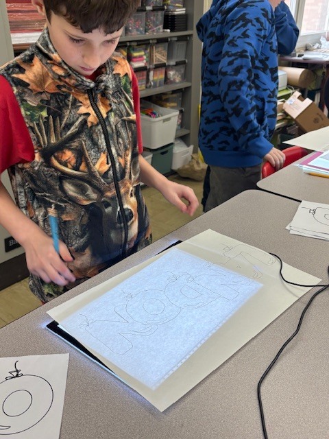 Students are tracing bubble letters using a light board. Their paper and the letter they are tracing on top of a board that is electronically lit up.