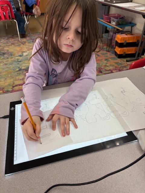 Students are tracing bubble letters using a light board. Their paper and the letter they are tracing on top of a board that is electronically lit up.