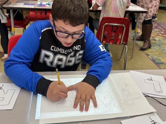 Students are tracing bubble letters using a light board. Their paper and the letter they are tracing on top of a board that is electronically lit up.