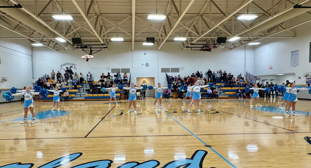 The cheerleaders are performing the school song with the band and crowd in the background. The cheerleaders are wearing school oolors of blue and yellow and are holding poms.