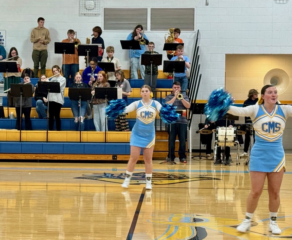 The band is standing in the bleachers playing the school song. Their stands are in front of them. Also pictured are two cheerleaders perfoming the school song.