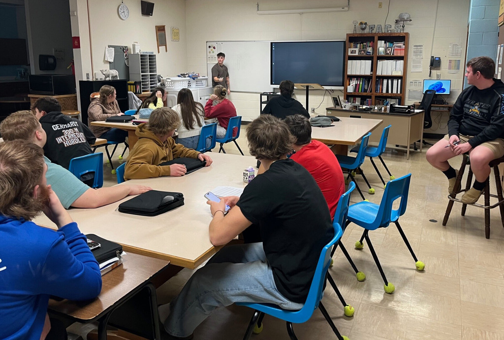 A student is standing at the white board. The student is drawing an object on the board. The other students are sitting at the tables and trying to guess what the drawing is. 