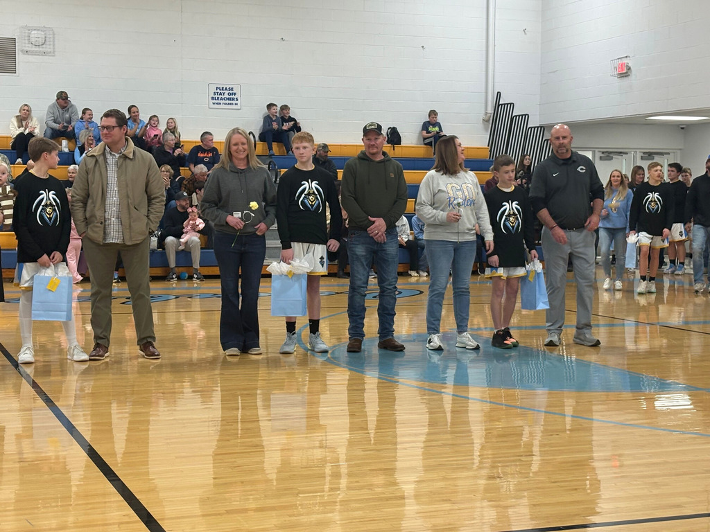 Parents and players from the basketball and cheer teams are standing in the center of the basketball court. Players are holding gift bags and parents are holding flowers. 