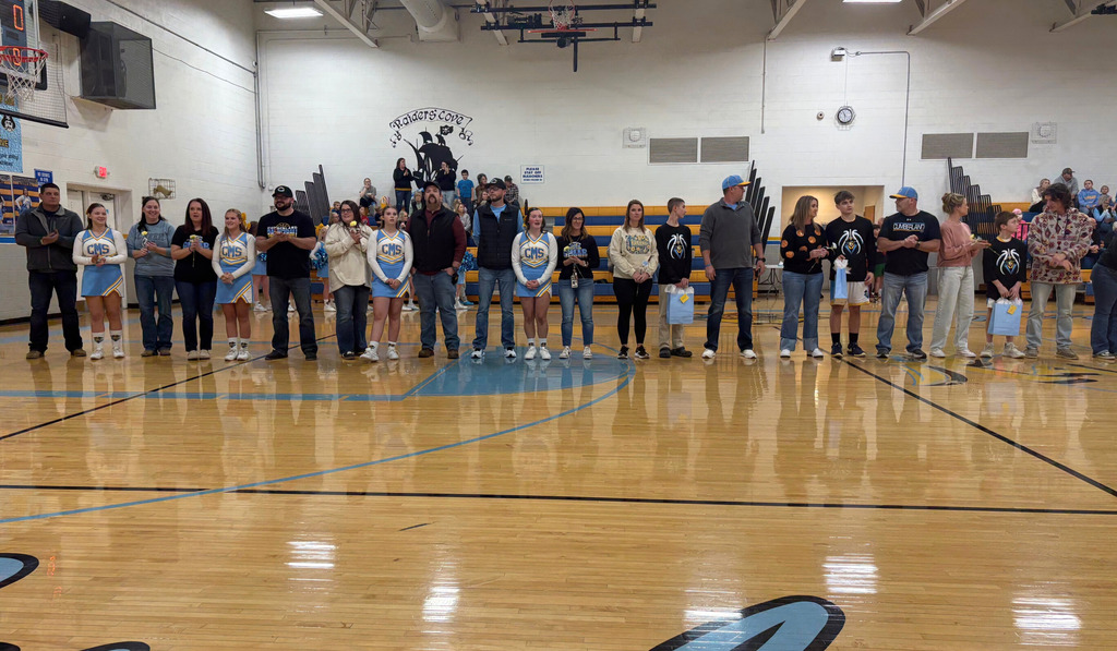 Parents and players from the basketball and cheer teams are standing in the center of the basketball court. Players are holding gift bags and parents are holding flowers. 