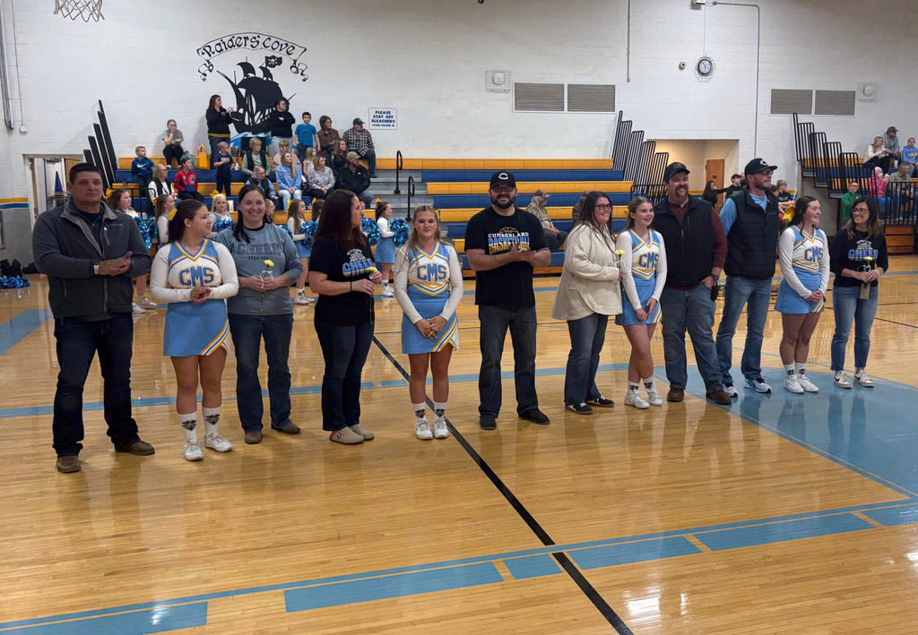 Parents and players from the basketball and cheer teams are standing in the center of the basketball court. Players are holding gift bags and parents are holding flowers. 
