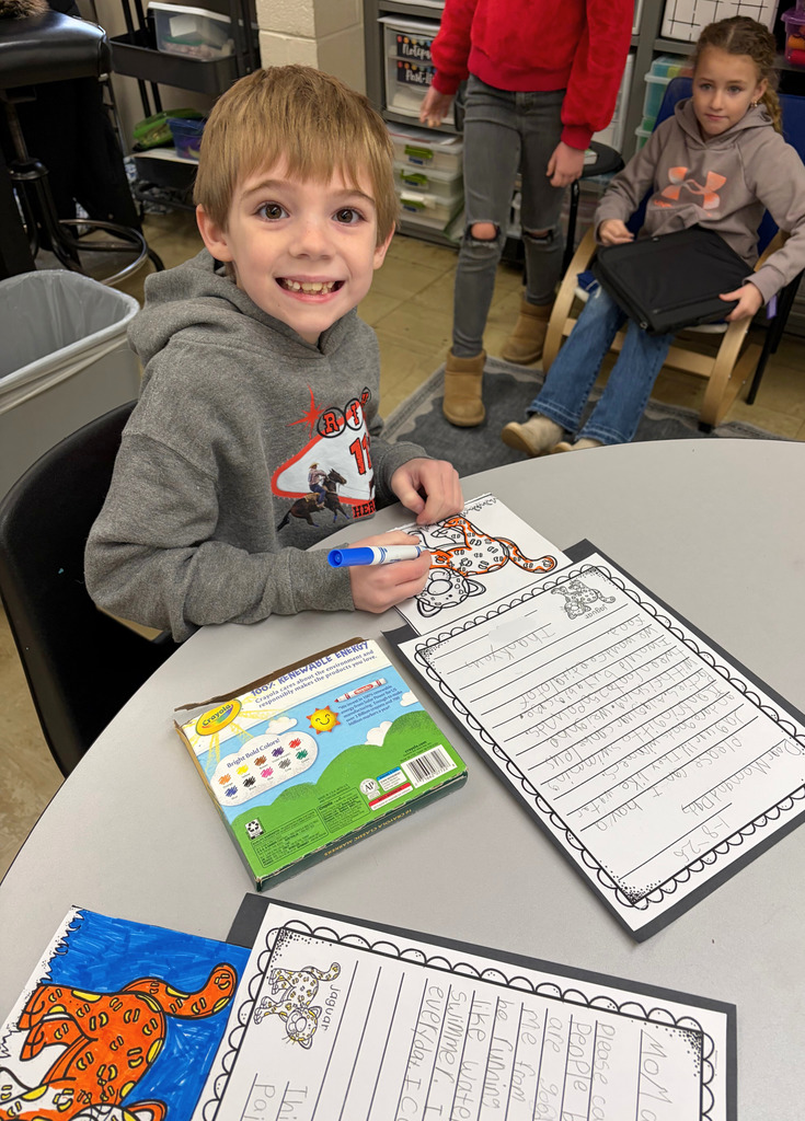 A student is sitting at his table. He is coloring a picture of a tiger to add to his letter. 