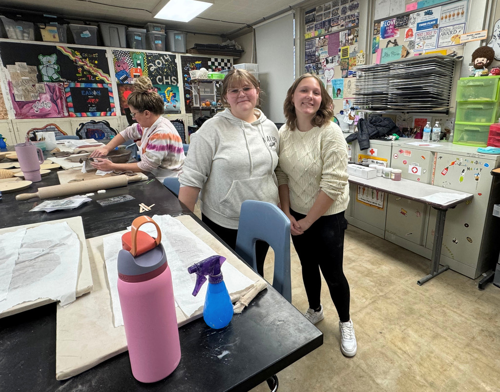Two student helpers are standing in the foreground. A participant is sitting in the background working on her clay bowl. 