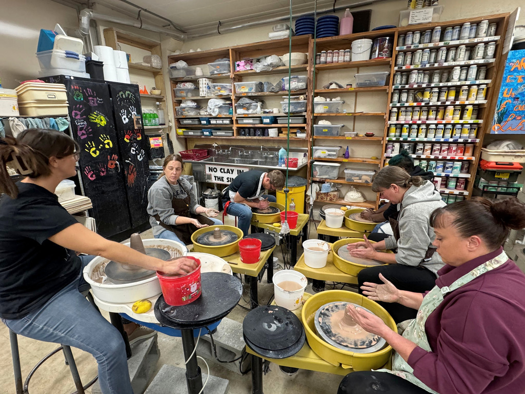 Six people are sitting at the wheels. They are manipulating their clay to get it to the correct form. 