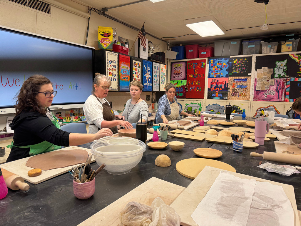 Three people are standing at the tables. They are working on clay projects. Another person is assisting each person.