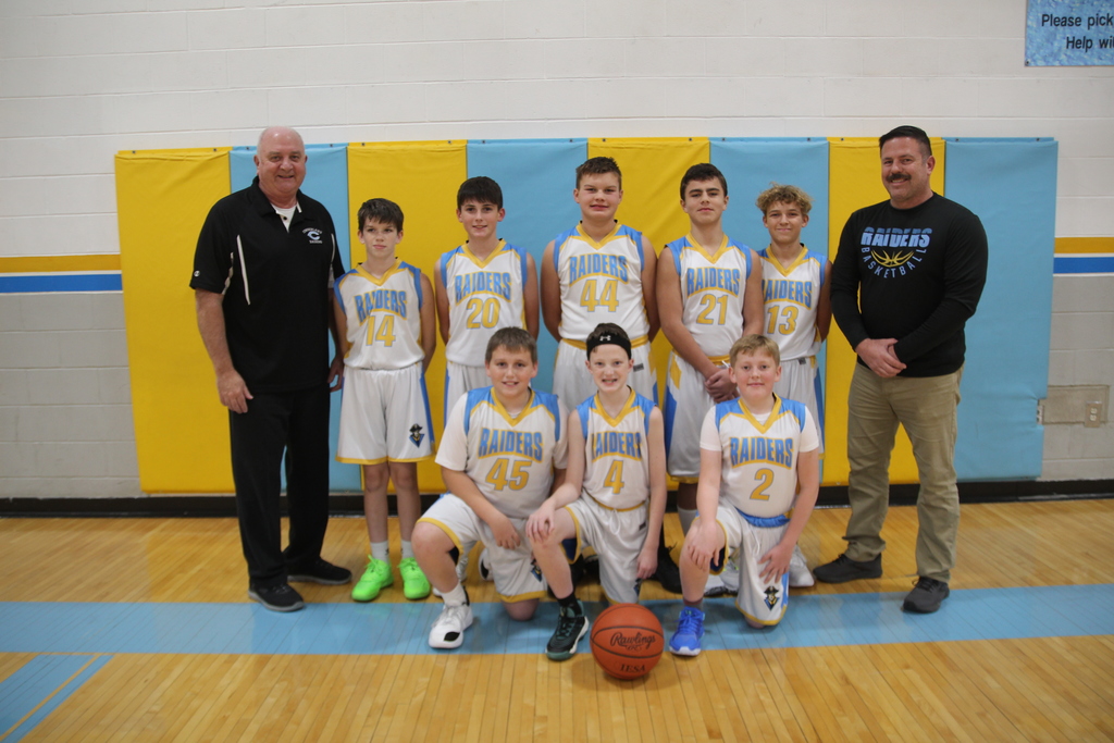 Pictured is teh seventh grade basketball team. The coaches are on each side of the team. There is a basketball on the floor in front of the team. 