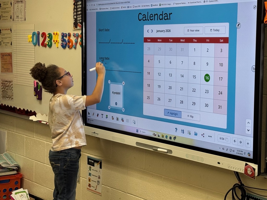 A student is standing at the Smartboard. The student is working on writing the date to the calendar. 