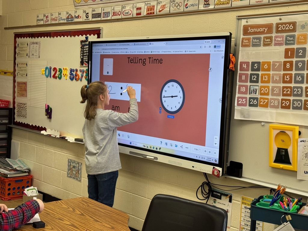 A student is standing at the Smartboard. The student is working on writing the time from a clock. 