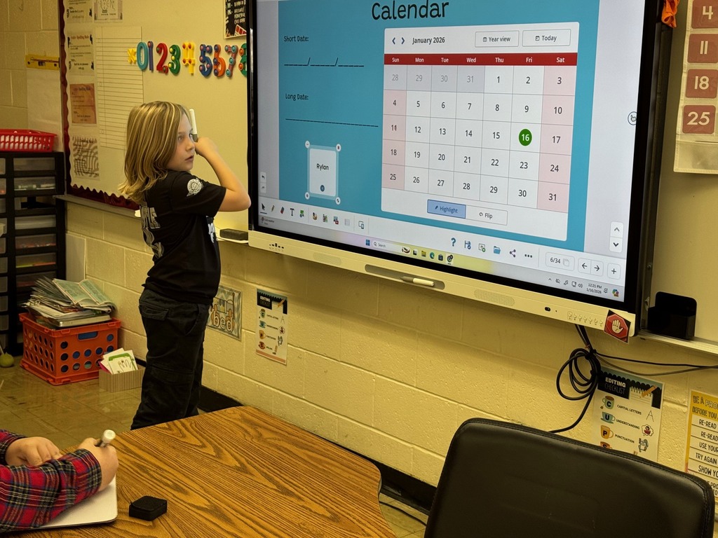 A student is standing at the Smartboard. The student is working on writing the date to the calendar. 