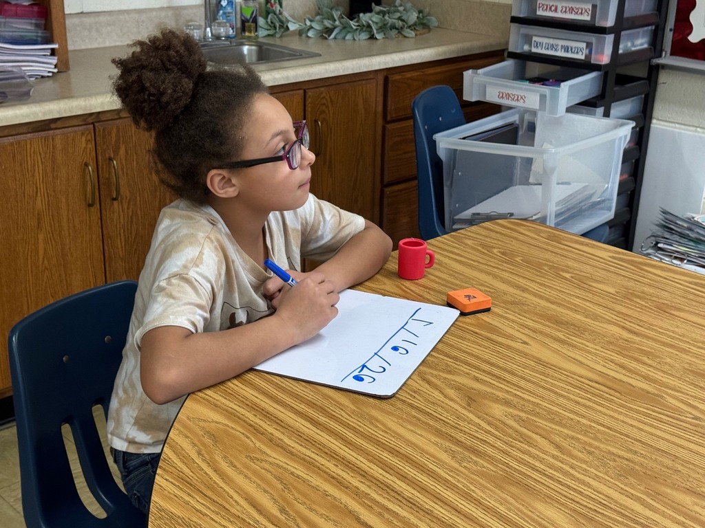 A student is sitting at a table. The student is working a problem on the white board. 