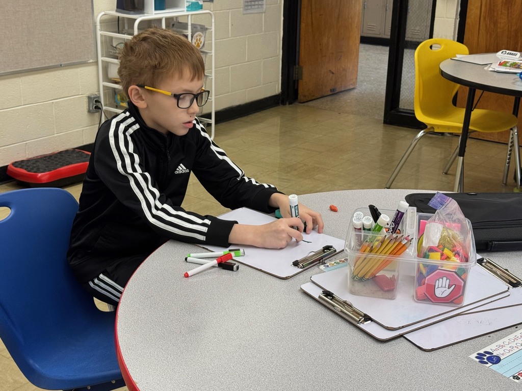 A student is sitting at a table. The student is working a problem on the white board. 