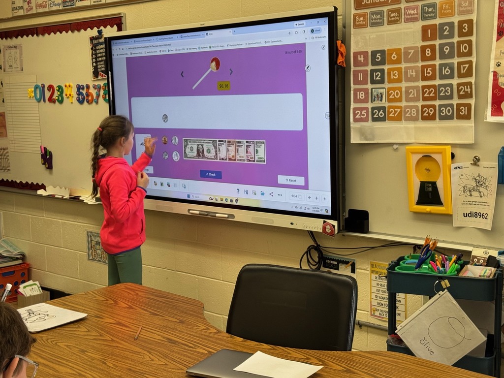 A student is standing at the Smartboard. The student is working on choosing the coins to pay for an intem.. 