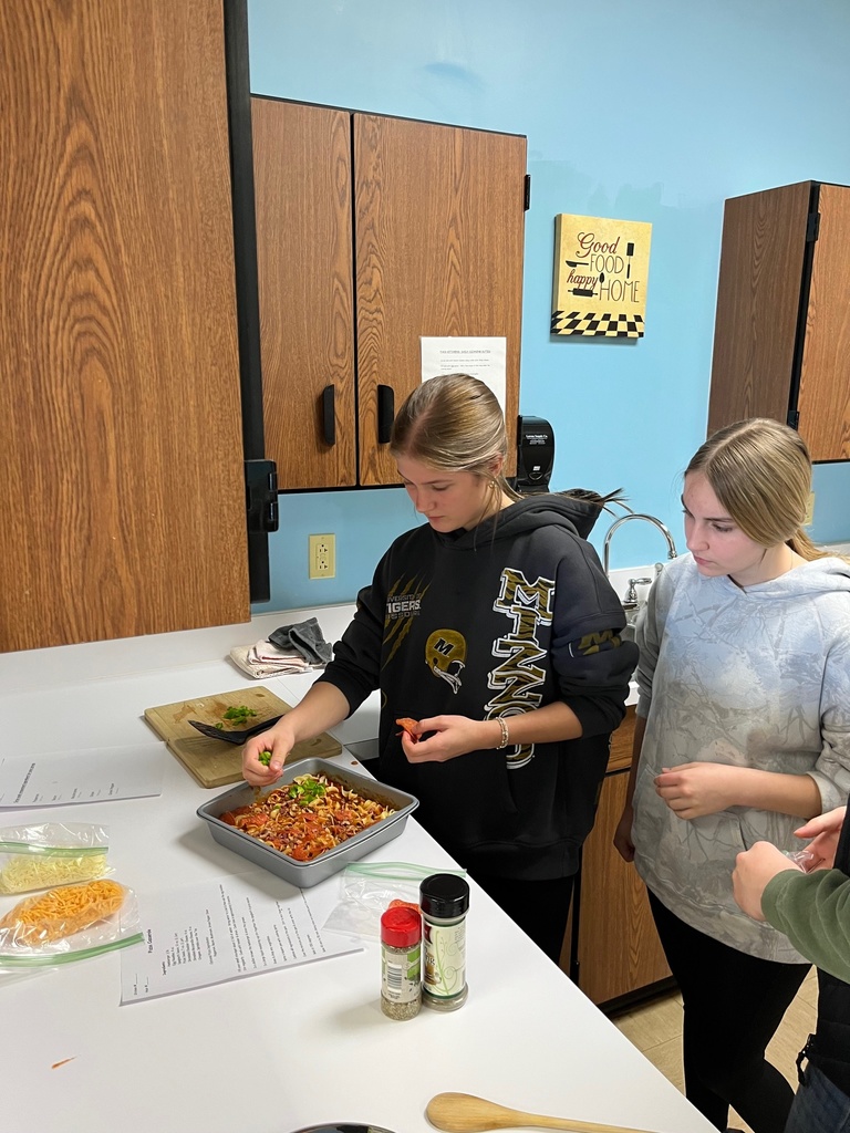 Students are at the counter. They are adding toppings to their pizza casserole. 