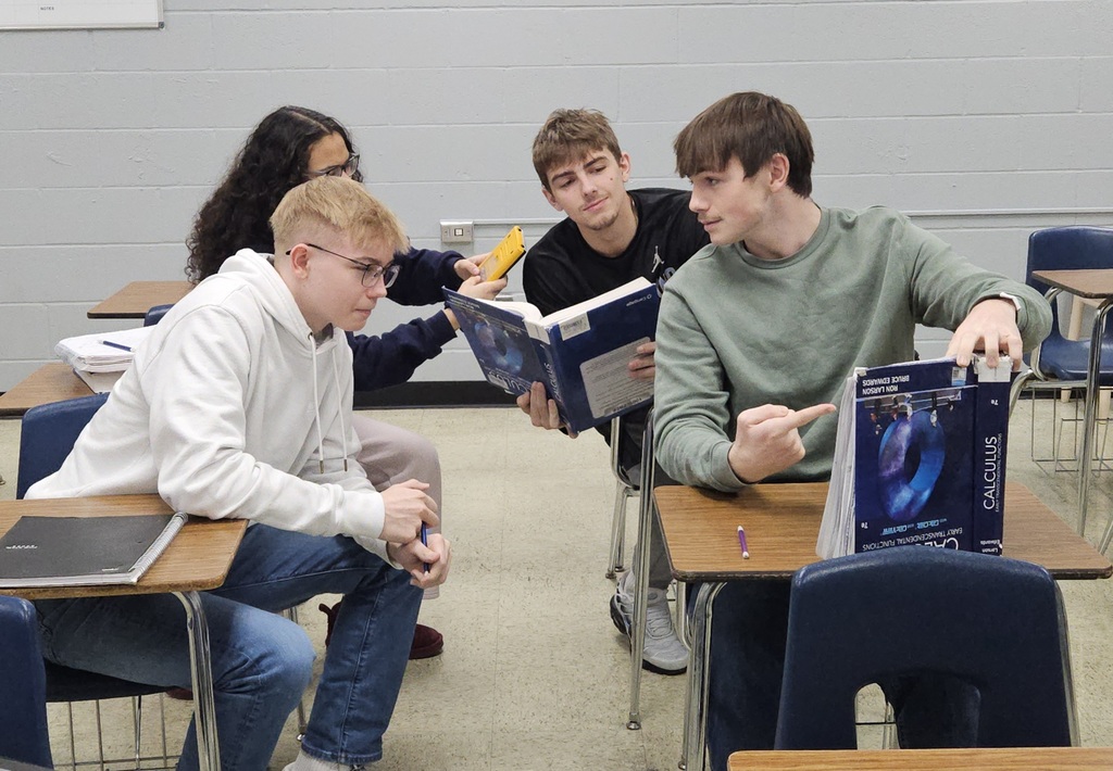 The four students are sitting in their desks. They are holding their Calculus books and calculators upside. down. 