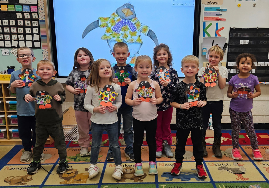 The students are standing in front of the smartboard which has a picture of Tacky the Penguin. The students are holding their penguins with their creative  Hawaiian shirts. 