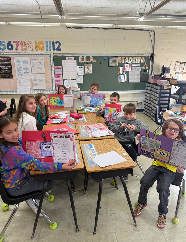 The students are sitting at their desks. They are each holding up their Love bug crafts. 