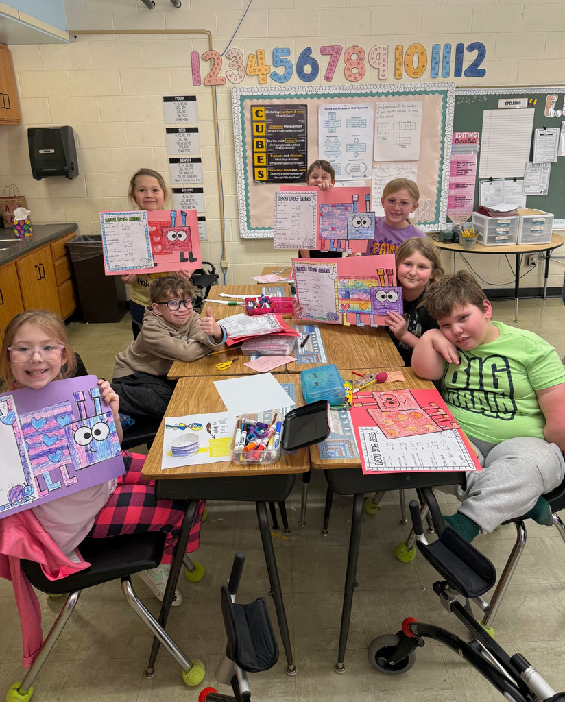 The students are sitting at their desks. They are each holding up their Love bug crafts. 