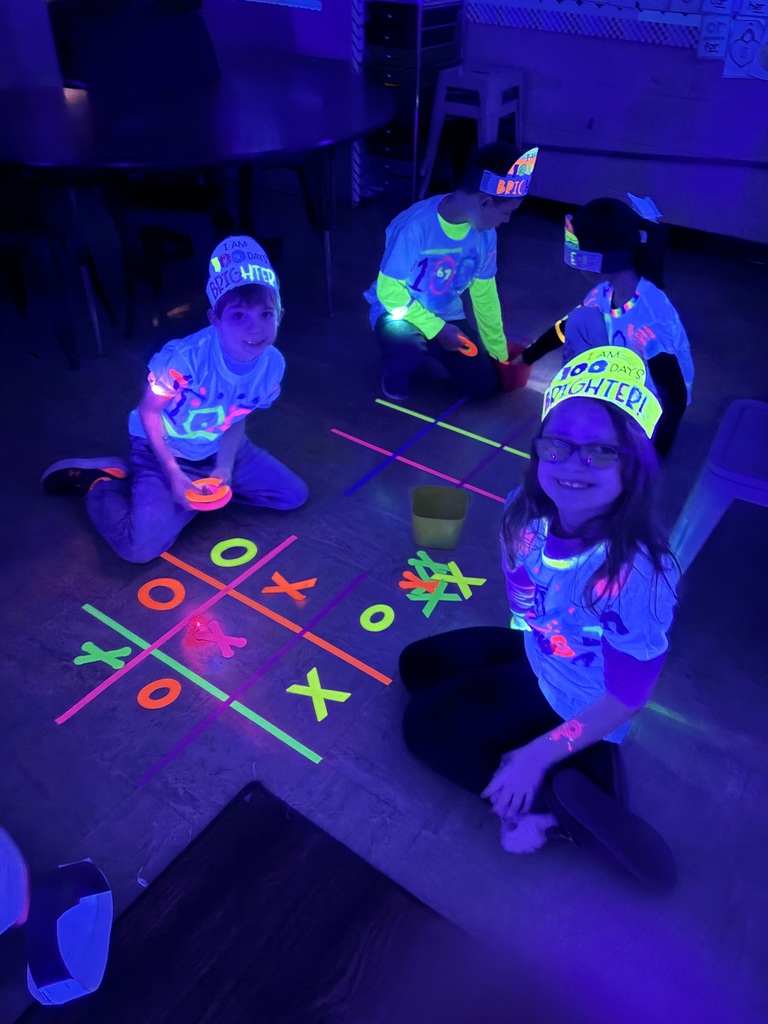 Students are sitting on the rug in the dark. They are playing glow-in-the-dark tik tac toe.