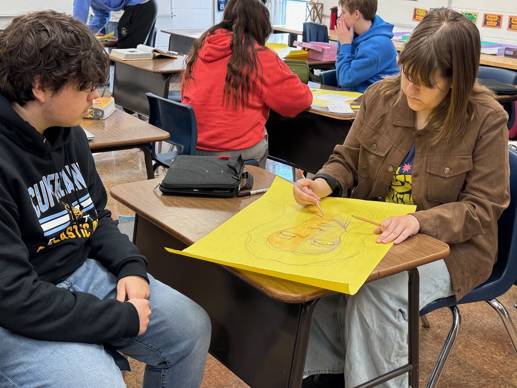 Students, working in groups are drawing one of the characters. The students are adding elements to the face. One side is what they show the world. The other side is what they hide. 