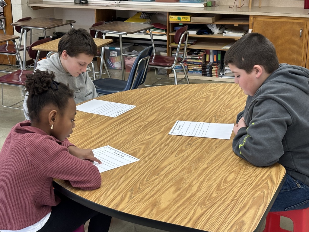 Three students are sitting at a kidney table. They are reading a passage aloud to each other. 