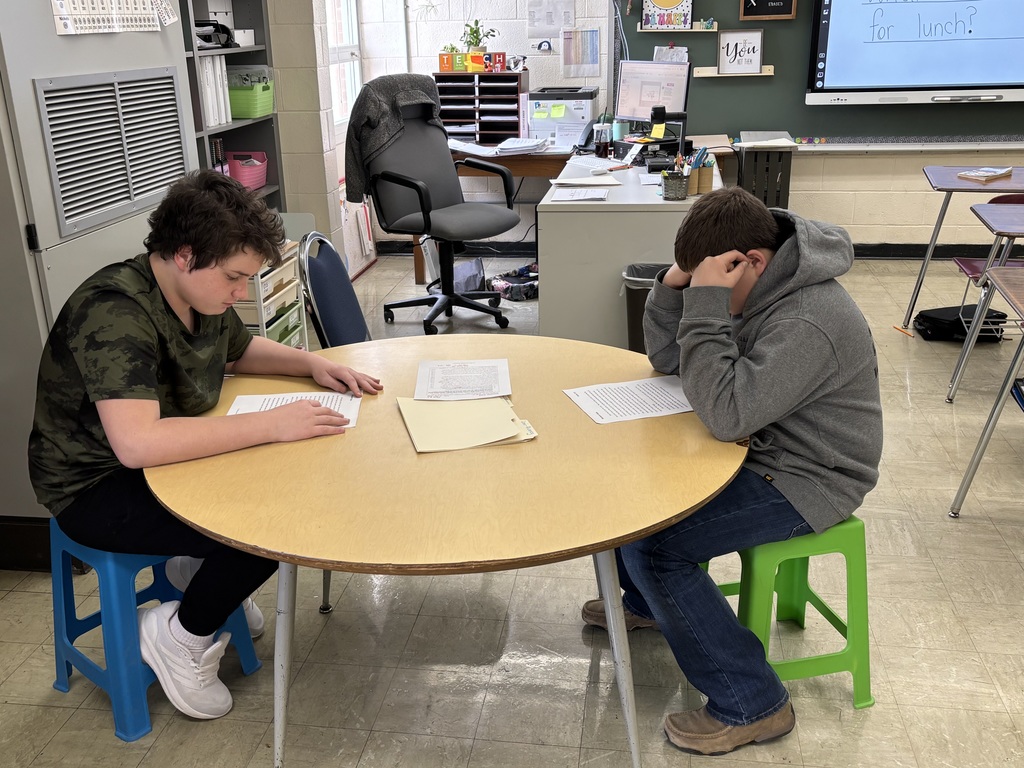Two students are sitting at a round table. They are reading a passage aloud to each other. 