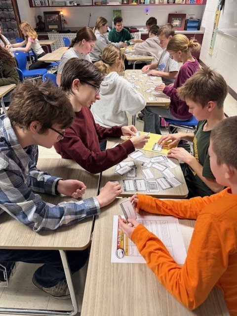 A group of four students are sitting with their desks facing each other. They have a document and a set  of cards on the desks. They are sorting the cards. 