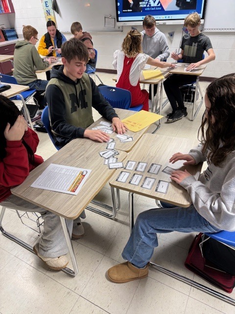 A group of three students are sitting with their desks facing each other. They have a document and a set  of cards on the desks. They are sorting the cards. 