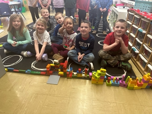 Seven students are standing behind the castle they made from building blocks. The castle has many blocks, turrets, and windows. 