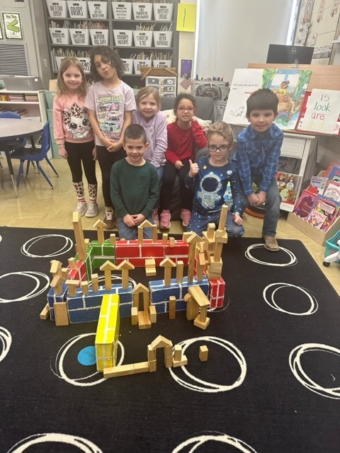 Seven students are standing behind the castle they made from building blocks. The castle has many blocks, turrets, and windows. 