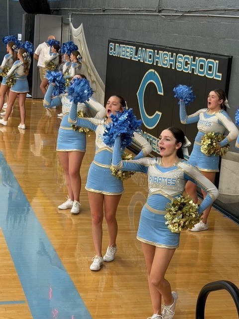 The cheerleaders are on the baseline. They have poms. They are cheering and have one pom in the air. 