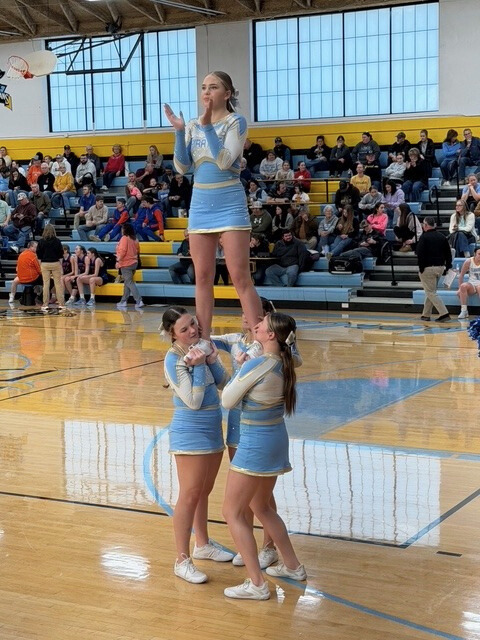 The cheerleaders are in a four person stunt. One cheerleader is lifted  up and clapping. The others are supporting her legs. 
