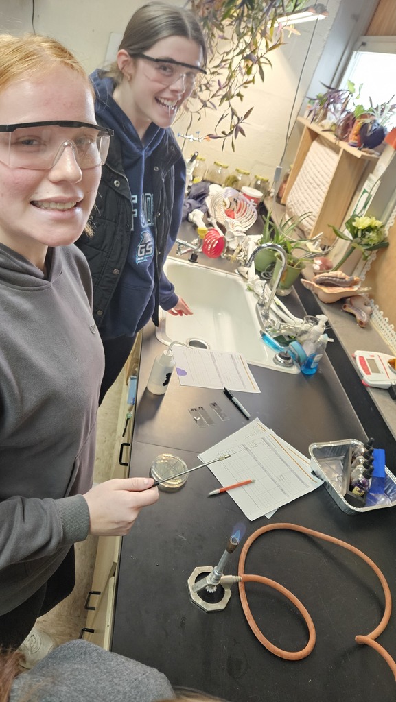 Students are standing at a lab table. One is holding a metal object over a bunsen burner and the other is preparing a petri dish. 