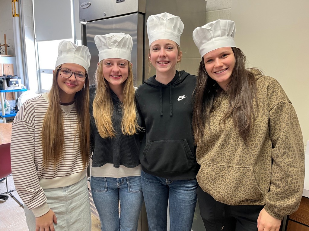 Four students are standing side by side. Each is wearing a chef hat. 
