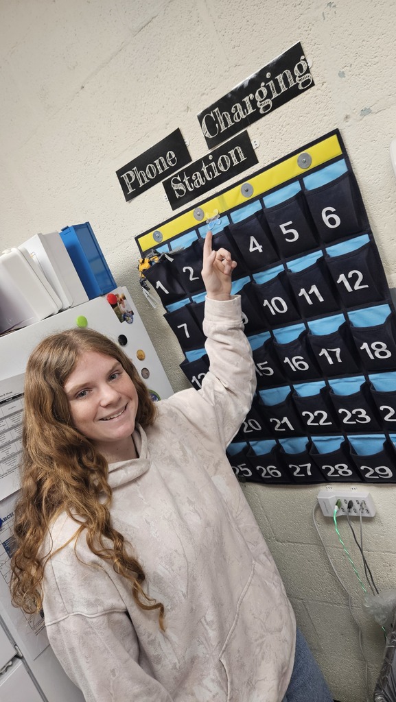 A student is pointing to the student's hidden butterfly. It is blue, yellow, and black blending in to the phone station. 