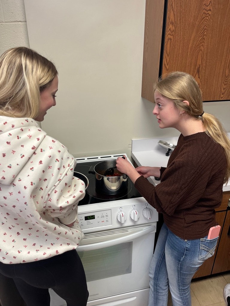 Two students are discussing their chilli recipe. They are standing over their pot at the stove.