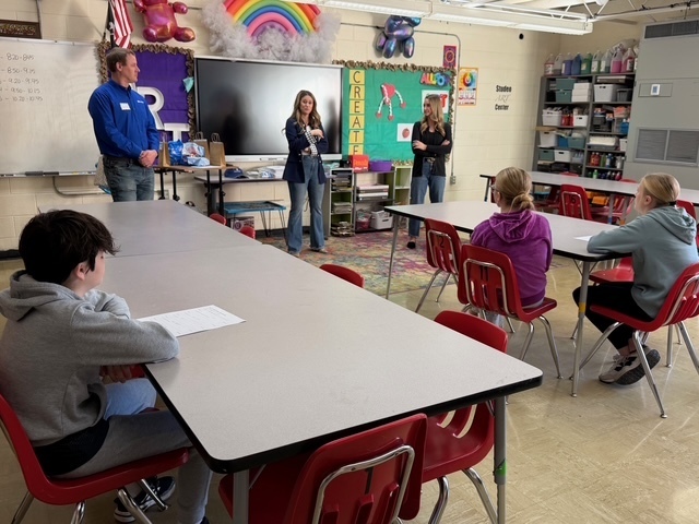 Three speakers are discussing his career with a group of students sitting in their desks.. 