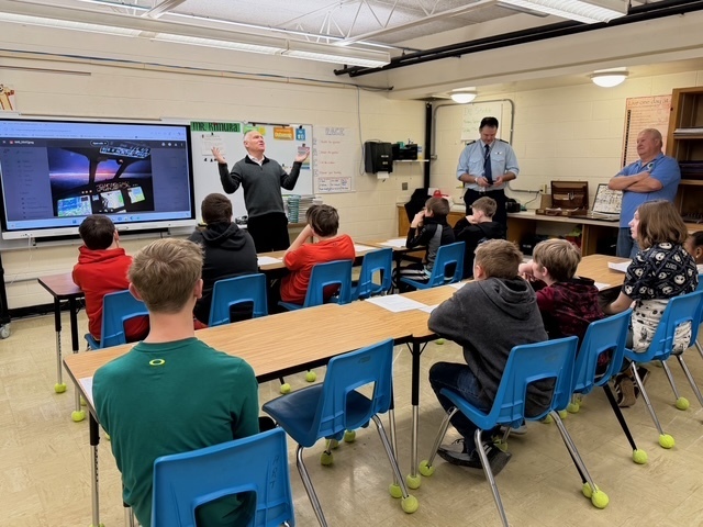 Three speakers are discussing his career with a group of students sitting in their desks.. There is a presentation on the screen. 