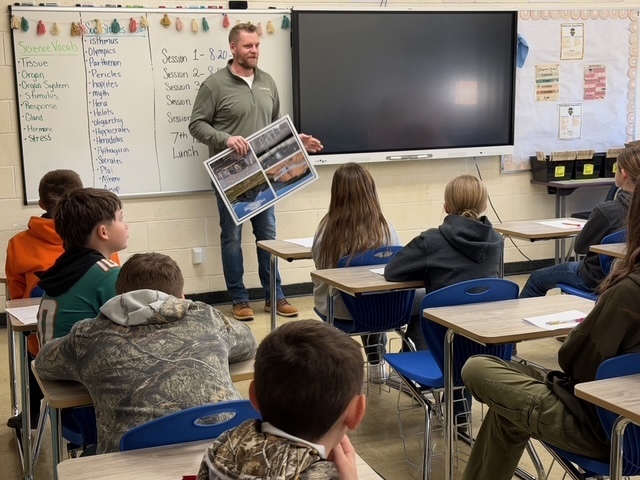 A speaker is discussing his career with a group of students sitting in their desks. He is holding a poster board. 