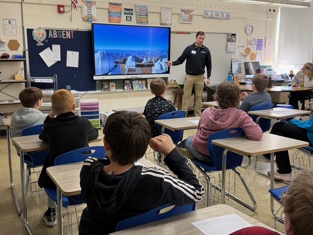 A speaker is discussing his career with a group of students sitting in their desks. There is a presentation on the screen.