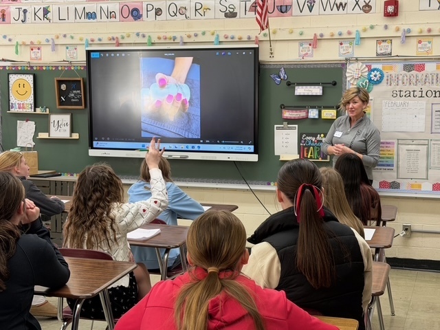 A speaker is discussing her career with a group of students sitting in their desks. There is a presentation on the screen.