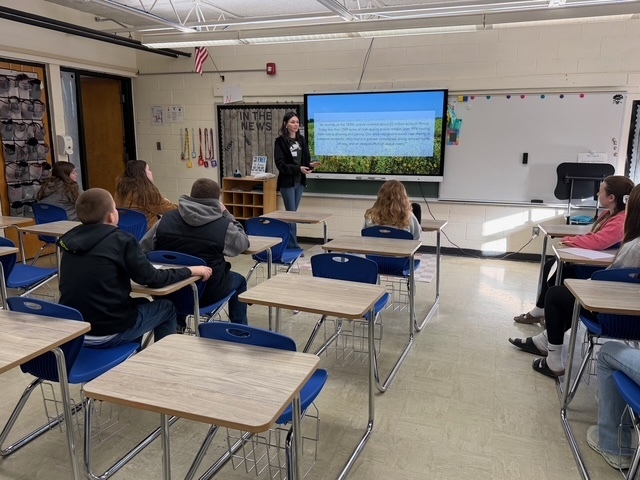 A speaker is discussing her career with a group of students sitting in their desks. There is a presentation on the screen.